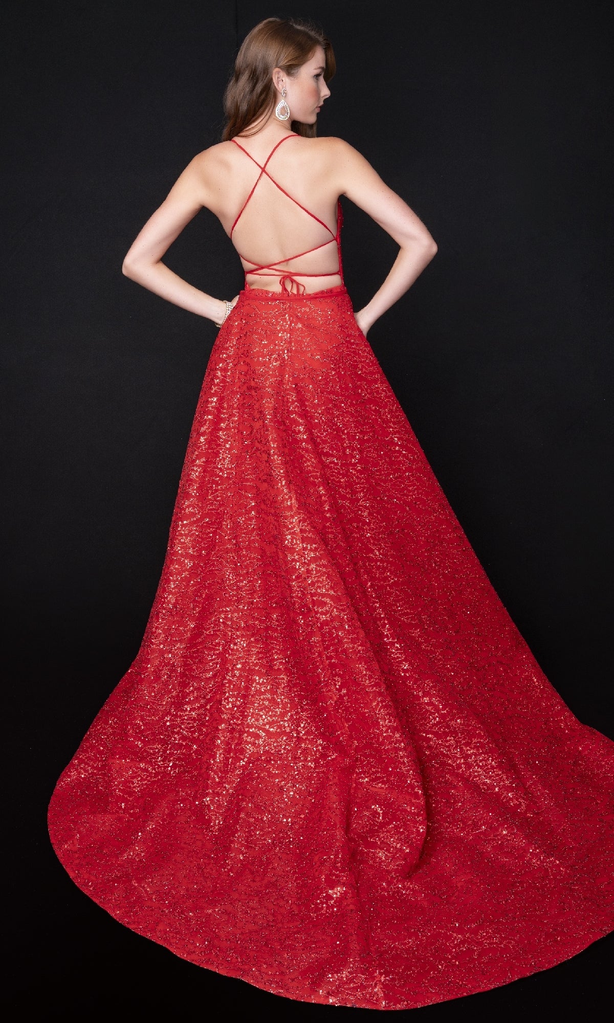 A woman faces away from the camera in a Nina Canacci 8250 long formal dress, showcasing its floor-length glitter design, strappy open back, and fitted bodice against a black background.